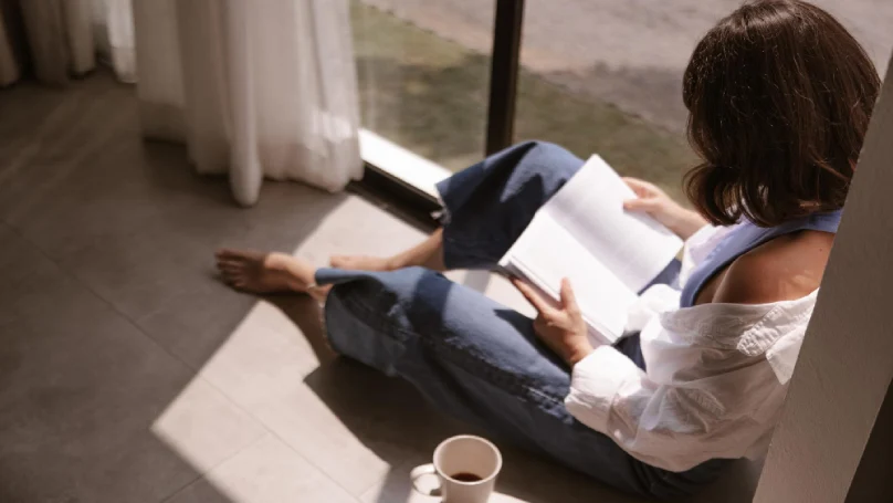 A person sitting by a window, deeply engrossed in reading a book, with a cup of coffee nearby.